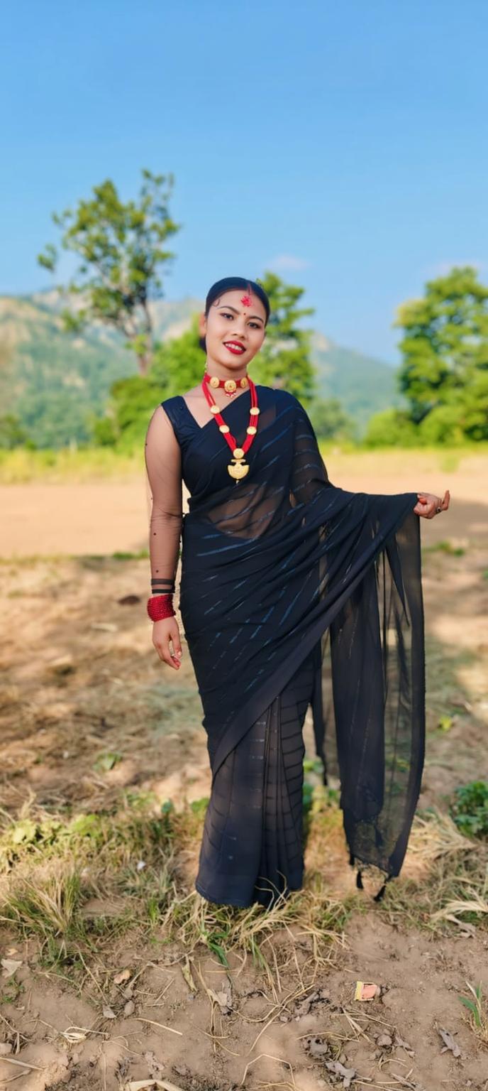 A beautiful woman in a traditional black saree, adorned with red accessories, standing in a serene outdoor location with mountains in the background and bright blue sky